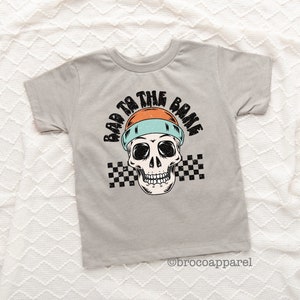 May include: A light gray short-sleeve t-shirt featuring a graphic of a skull wearing an orange and light blue beanie. The text "BAD TO THE BONE" is printed in black above the skull, with a black and white checkered pattern behind the skull.