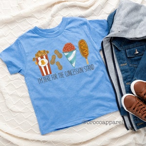 May include: A light blue t-shirt featuring a graphic of popcorn, peanuts, a snow cone, and a corn dog. The text "I'm here for the concession stand" is printed on the shirt.