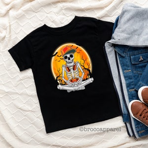 May include: Black t-shirt featuring a skeleton wearing a cap and headphones, set against an orange moon backdrop with bats and trees. The skeleton is in a seated position. A denim jacket, gray hoodie, and brown sneakers are next to the shirt. The text "brocoapparel" is at the bottom.