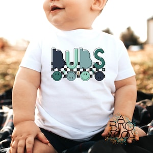 May include: White short-sleeved t-shirt with the word "BUBS" in blue and green letters. Below the word are four smiley faces with lightning bolts on a black and white checkered background. The shirt is for babies.