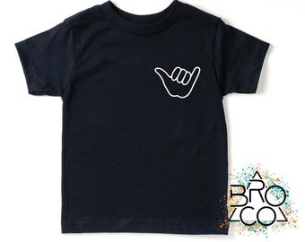 May include: A black short-sleeve t-shirt featuring a white outline of a shaka hand gesture on the left chest area. The shirt has a crew neckline and a straight hem. A logo with the text BRO CO is visible in the bottom right corner of the image.