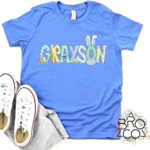May include: A blue t-shirt with the name "GRAYSON" spelled out in colorful letters with Easter-themed designs. The letters are decorated with bunnies, carrots, and eggs.