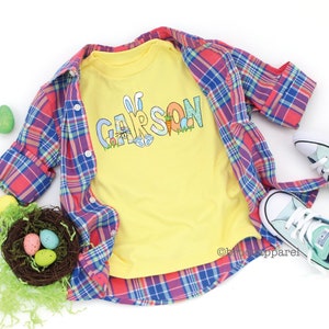 May include: A yellow t-shirt with the name "CARSON" spelled out in colorful letters decorated with Easter-themed graphics, such as bunnies, eggs, and carrots. The shirt is laid on a plaid shirt and surrounded by Easter eggs.