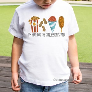 May include: White t-shirt featuring a graphic of popcorn, peanuts, a snow cone, and a corn dog. The text on the shirt reads "I'm here for the concession stand."