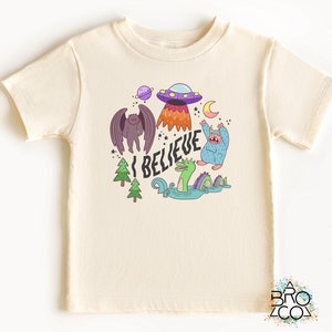 May include: Cream-colored t-shirt featuring a colorful graphic with the text "I BELIEVE". The graphic includes a purple UFO, a winged creature, a Bigfoot-like monster, a Loch Ness Monster, and other whimsical elements. A fun, playful design.