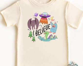Puede incluir: Camiseta de color crema con un gráfico colorido y el texto "I BELIEVE". El gráfico incluye un OVNI morado, una criatura alada, un monstruo parecido a Bigfoot, un monstruo del lago Ness y otros elementos caprichosos. Un diseño divertido y juguetón.
