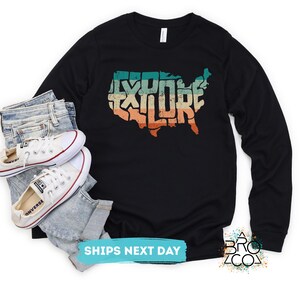 May include: A black long-sleeve shirt featuring a graphic of the United States map with the word EXPLORE written inside in a distressed, multi-colored retro font, paired with light blue denim jeans and white canvas sneakers.