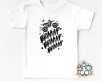 May include: A white short-sleeve t-shirt featuring a black graphic of a dirt bike and the repeated text BRAAAP. The design includes scattered black dots and motion lines surrounding the motorcycle, creating a dynamic visual effect on the chest.