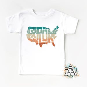 May include: A white short-sleeve t-shirt featuring a graphic of the contiguous United States map filled with the word EXPLORE in a distressed, multi-colored horizontal striped pattern of teal, beige, and orange. A logo reads BRO CO.