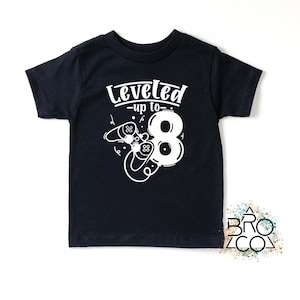 May include: A navy blue t-shirt featuring the text "Leveled up to 8" in white, along with a number 8 and game controller graphics. This t-shirt is designed for children.