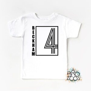 May include: A white short-sleeved t-shirt featuring a black graphic design on the chest. The design consists of the word BECKHAM written vertically in a sans-serif font next to a large number 4 enclosed within a black rectangular frame.