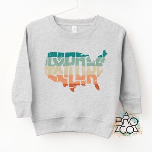 May include: A light gray long-sleeve sweatshirt featuring a distressed graphic of the United States map filled with horizontal stripes in teal, beige, and orange. The word EXPLORE is integrated into the map design in a bold, blocky font.
