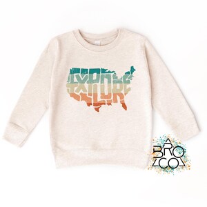 May include: A heathered beige long-sleeve sweatshirt featuring a graphic of the United States map filled with the word EXPLORE in horizontal stripes of teal, cream, and orange. The logo BRO CO is printed in the bottom right corner.
