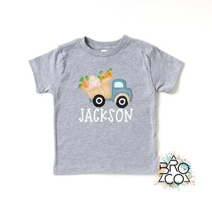 May include: A gray short-sleeved t-shirt with a blue truck carrying a basket of carrots and a bunny. The text "JACKSON" is printed below the truck.