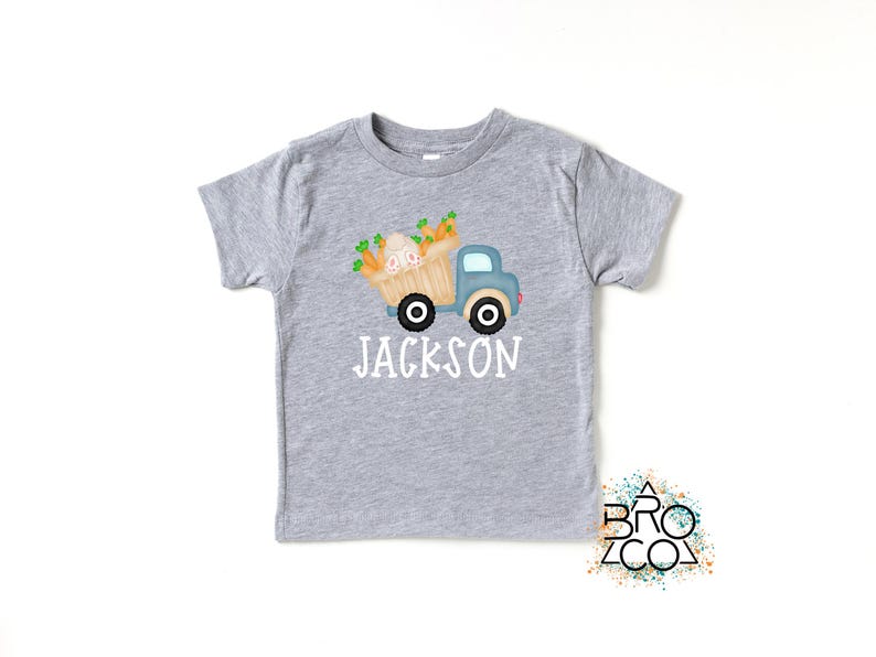 May include: A gray short-sleeved t-shirt with a blue truck carrying a basket of carrots and a bunny. The text "JACKSON" is printed below the truck.