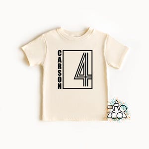 May include: A cream-colored short-sleeve t-shirt featuring a black graphic design on the front. The design consists of a rectangular frame containing the name CARSON written vertically and a large number 4 with a double-line outline style.