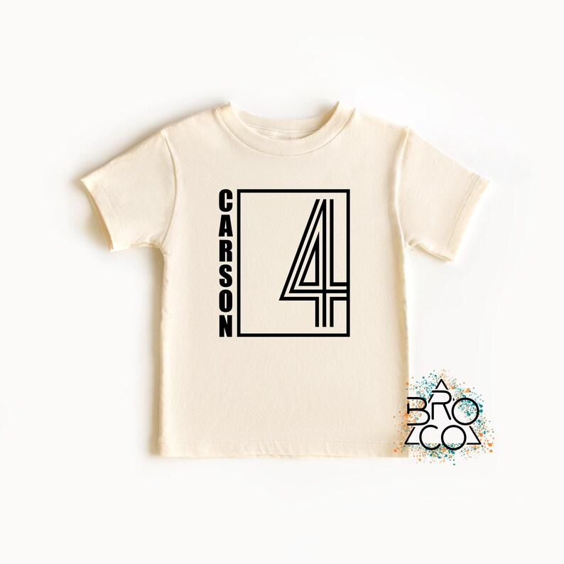 May include: A cream-colored short-sleeve t-shirt featuring a black graphic design on the front. The design consists of a rectangular frame containing the name CARSON written vertically and a large number 4 with a double-line outline style.