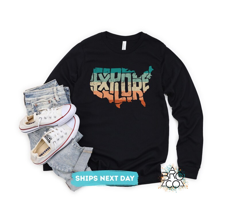May include: A black long-sleeve shirt featuring a graphic of the United States map with the word EXPLORE written inside in a distressed, multi-colored retro font, paired with light blue denim jeans and white canvas sneakers.