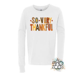 May include: A white long-sleeve shirt featuring the text SO VERY THANKFUL in a multi-colored, stylized font with shades of orange, brown, and yellow. The design is centered on the chest, and a small logo with the text BRO CO is visible at the bottom right.
