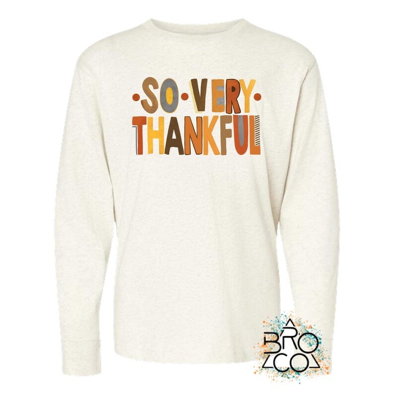 May include: A cream-colored long-sleeve shirt featuring the text SO VERY THANKFUL printed in a multi-colored, block-style font with shades of brown, orange, and yellow. The shirt has a crew neckline and a heathered fabric texture throughout.