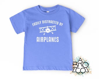 May include: A light blue short-sleeve t-shirt featuring a white graphic of a biplane and the text "EASILY DISTRACTED BY AIRPLANES" printed in white across the chest. The shirt has a crew neckline and a standard fit for casual wear.
