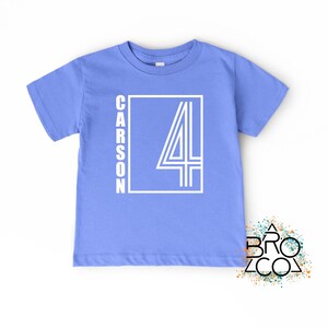 May include: A light blue short-sleeved t-shirt featuring a white graphic design on the front. The design consists of a rectangular frame containing the name CARSON written vertically and a large number 4 with a triple-line outline style.