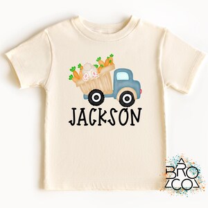 May include: A light beige t-shirt with a blue truck carrying a basket of carrots and a bunny. The text "JACKSON" is printed below the truck.