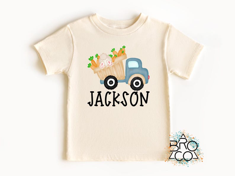 May include: A light beige t-shirt with a blue truck carrying a basket of carrots and a bunny. The text "JACKSON" is printed below the truck.