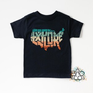 May include: A black short-sleeve t-shirt featuring a graphic of the United States map filled with the word EXPLORE in a distressed, multi-colored horizontal striped pattern of teal, beige, and orange. The design has a weathered texture.