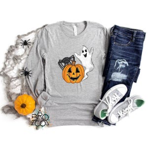 May include: A gray long-sleeve shirt featuring a Halloween-themed graphic. The design includes a smiling orange pumpkin, a black spider, and a white ghost. The shirt is paired with blue jeans and gray sneakers.