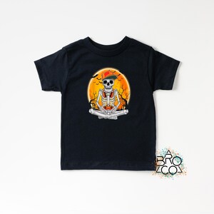 May include: Black t-shirt featuring a skeleton wearing a hat and headphones, sitting in a meditative pose with a video game controller. The design includes an orange moon background and trees, perfect for Halloween or gaming enthusiasts.