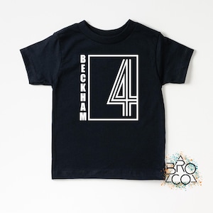 May include: A black short-sleeved t-shirt featuring a white graphic print on the chest. The design includes the word BECKHAM written vertically in a sans-serif font next to a large white outlined number 4 inside a rectangular frame.