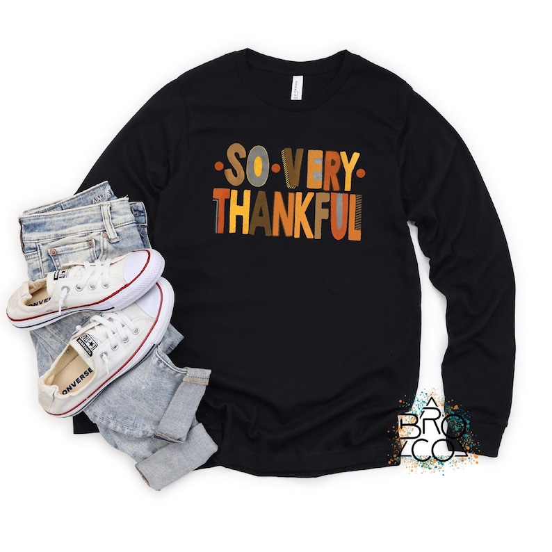 May include: A black long-sleeve t-shirt featuring the text SO VERY THANKFUL in multi-colored block letters, paired with light blue denim jeans and white canvas sneakers with white laces and rubber soles.