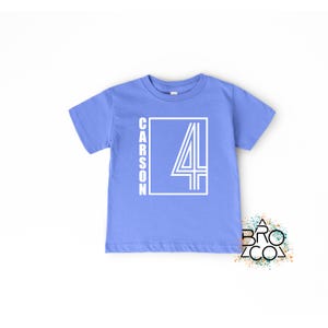 May include: A light blue short-sleeve t-shirt featuring a white graphic design on the front. The design consists of a rectangular frame containing the name CARSON written vertically and a large number 4 with a double-line outline style.