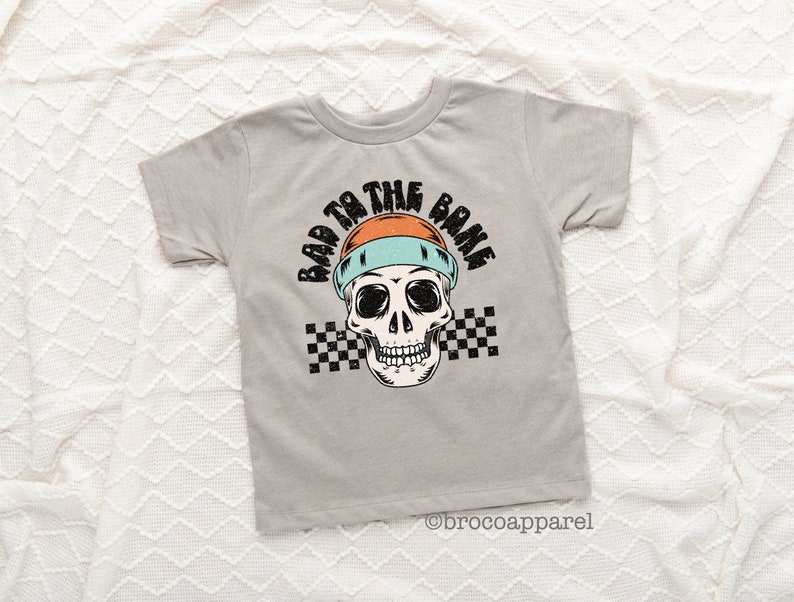 May include: A light gray short-sleeve t-shirt featuring a graphic of a skull wearing an orange and light blue beanie. The text "BAD TO THE BONE" is printed in black above the skull, with a black and white checkered pattern behind the skull.