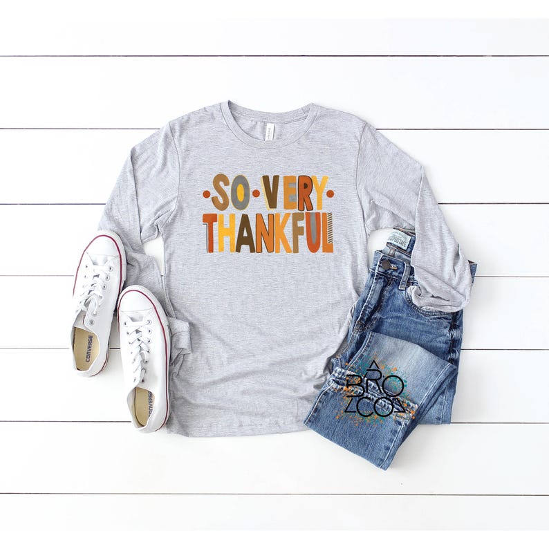 May include: A heather gray long-sleeve shirt with the text SO VERY THANKFUL in orange, brown, and yellow letters, paired with blue denim jeans and white canvas sneakers with white laces and rubber soles.