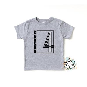 May include: A heather gray short-sleeve t-shirt featuring a black graphic design on the front. The design includes the name CARSON written vertically next to a large outlined number 4 inside a rectangular frame. A small logo reads BRO CO.