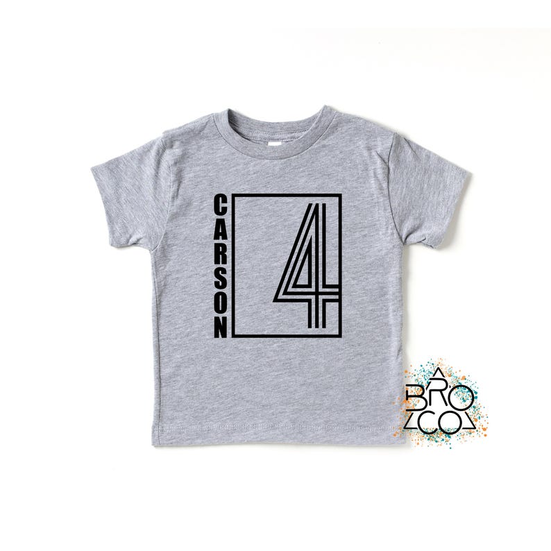 May include: A heather gray short-sleeve t-shirt featuring a black graphic design on the front. The design includes the name CARSON written vertically next to a large outlined number 4 inside a rectangular frame. A small logo reads BRO CO.