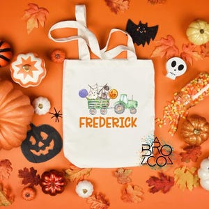 Puede incluir: Una bolsa de mano de lona blanca con un dise&ntilde;o tem&aacute;tico de Halloween que presenta un tractor verde tirando de un vag&oacute;n con fantasmas y calabazas. El nombre "Frederick" est&aacute; impreso en naranja. El fondo incluye calabazas, hojas y decoraciones de Halloween.