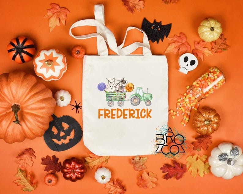 Puede incluir: Una bolsa de mano de lona blanca con un dise&ntilde;o tem&aacute;tico de Halloween que presenta un tractor verde tirando de un vag&oacute;n con fantasmas y calabazas. El nombre "Frederick" est&aacute; impreso en naranja. El fondo incluye calabazas, hojas y decoraciones de Halloween.