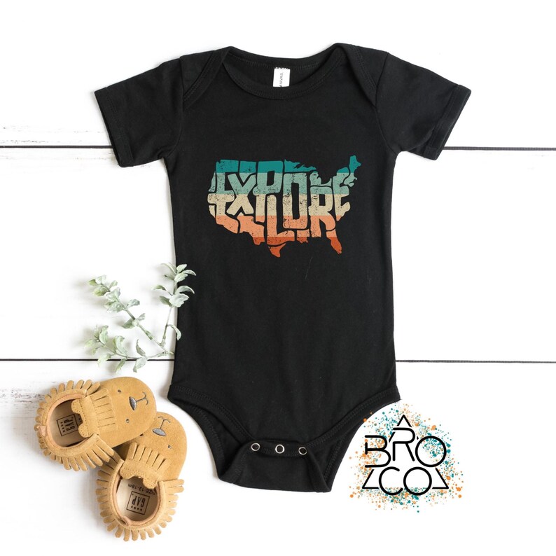 May include: A black short-sleeved baby bodysuit featuring a graphic of the United States map with the word EXPLORE integrated into the shape. The graphic uses a distressed, retro-style color palette of teal, cream, and orange tones.