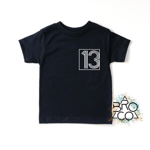 Op de afbeelding: Een marineblauw t-shirt met het nummer "13" in een witte, retro-stijl, omlijnd door een wit vierkant. Het t-shirt heeft een ronde hals en korte mouwen. Eenvoudig, stijlvol ontwerp.