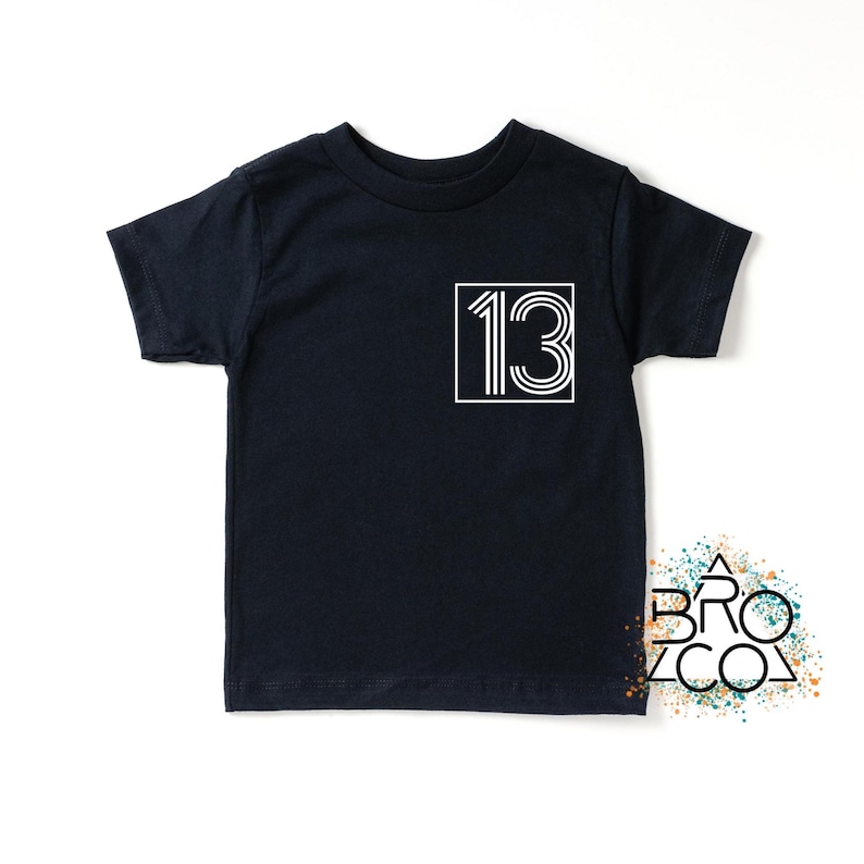 Op de afbeelding: Een marineblauw t-shirt met het nummer "13" in een witte, retro-stijl, omlijnd door een wit vierkant. Het t-shirt heeft een ronde hals en korte mouwen. Eenvoudig, stijlvol ontwerp.