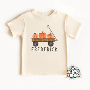 May include: A cream-colored short-sleeve t-shirt featuring a graphic of a brown wooden wagon filled with three orange pumpkins. The wagon has a silver handle and black wheels. The name FREDERICK is printed in black capital letters below the wagon.