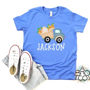 May include: A light blue t-shirt with a graphic of a blue truck carrying a basket of carrots and bunnies. The text "JACKSON" is printed in white below the truck.