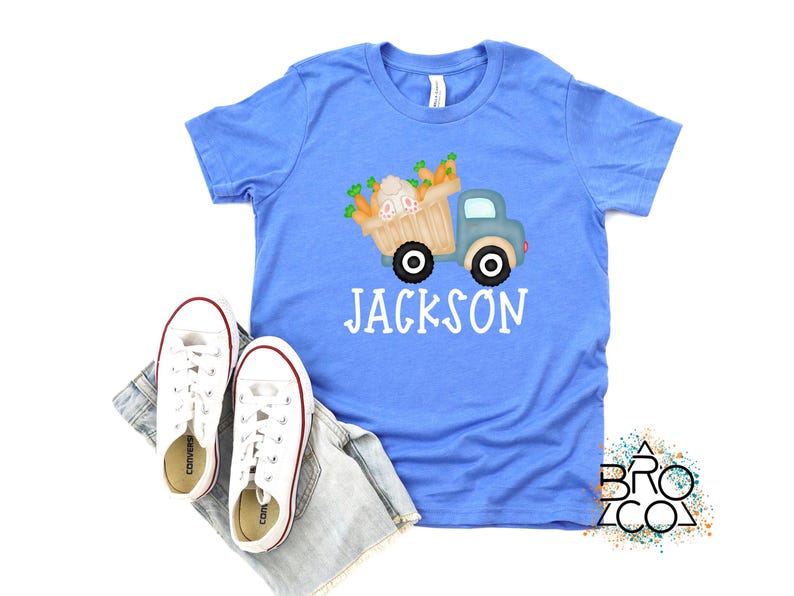 May include: A light blue t-shirt with a graphic of a blue truck carrying a basket of carrots and bunnies. The text "JACKSON" is printed in white below the truck.