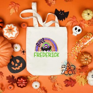 Puede incluir: Un bolso tote blanco con un dise&ntilde;o de arco&iacute;ris colorido, un murci&eacute;lago y el nombre "Frederick" en verde. La bolsa est&aacute; rodeada de decoraciones de Halloween, incluyendo calabazas, hojas y una calavera.