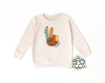 May include: Beige sweatshirt with a colorful turkey hand graphic. The turkey hand has a blue, orange, yellow, and brown design, with the word "Thankful" written below. The sweatshirt is a crew neck style.