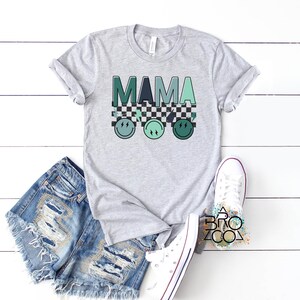 May include: A grey t-shirt with the word "MAMA" in green and blue letters. The letters are on top of a black and white chequered pattern. There are three green and blue smiley faces below the chequered pattern.