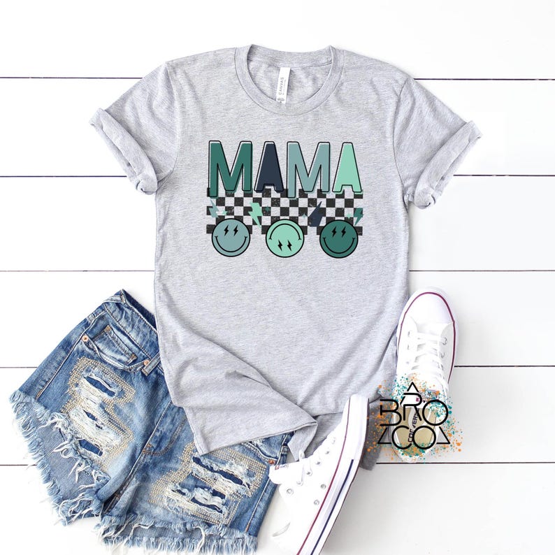 May include: A grey t-shirt with the word "MAMA" in green and blue letters. The letters are on top of a black and white chequered pattern. There are three green and blue smiley faces below the chequered pattern.